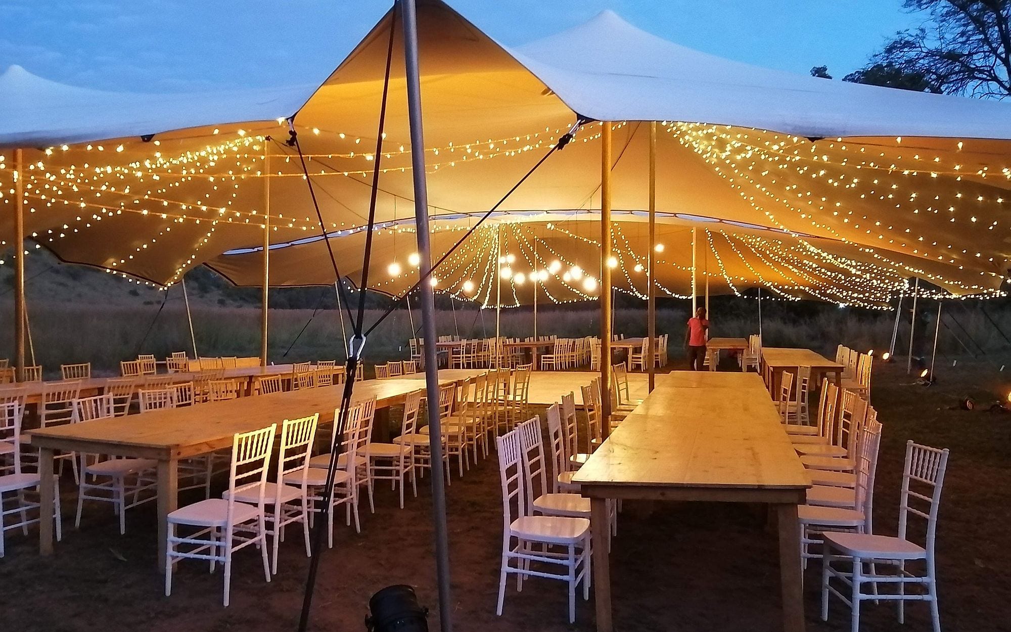 A stretch tent strung with fairy lights over long wooden tables at dusk